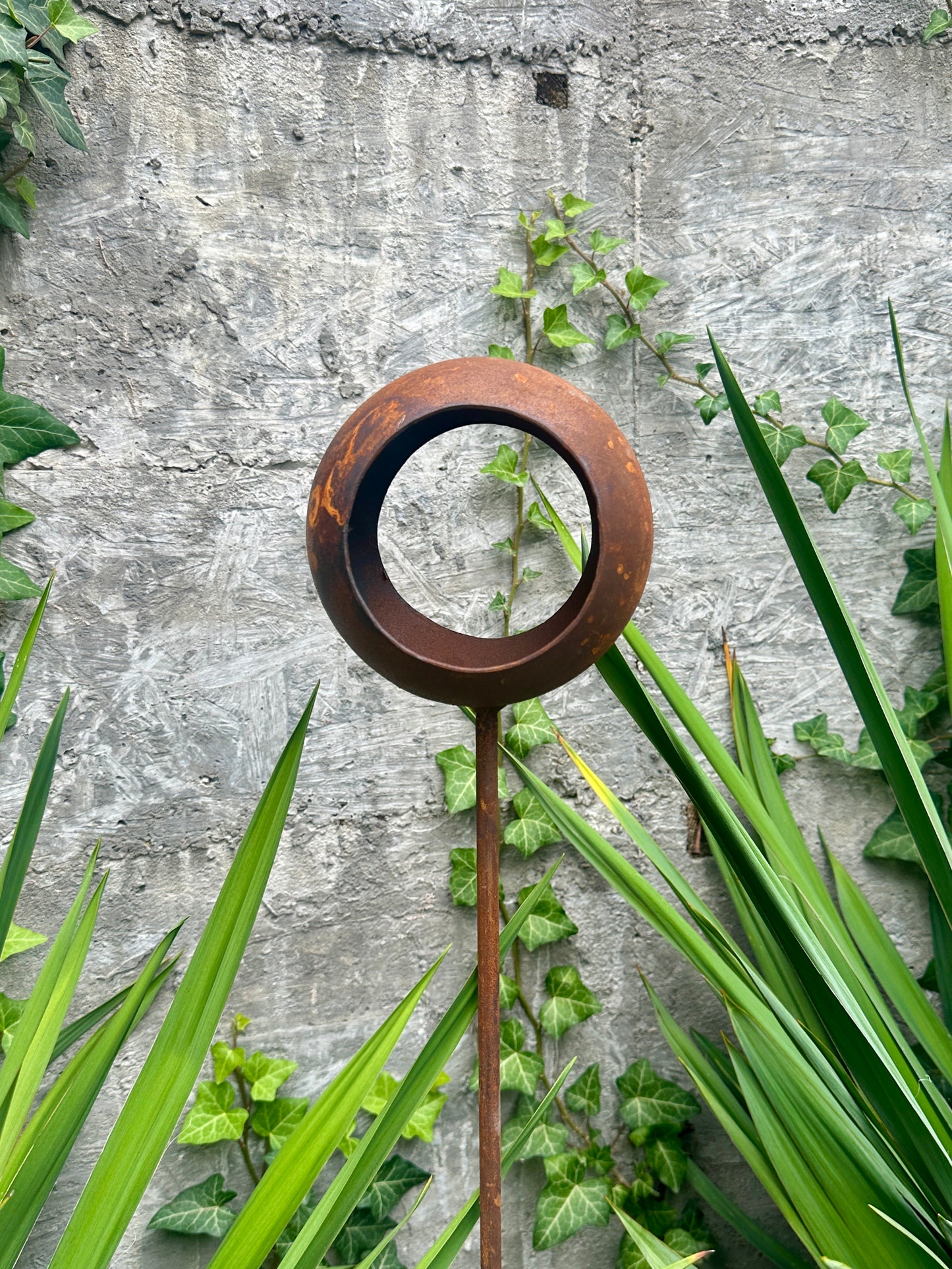 Decorative metal stake with circular design against a stone wall and greenery
