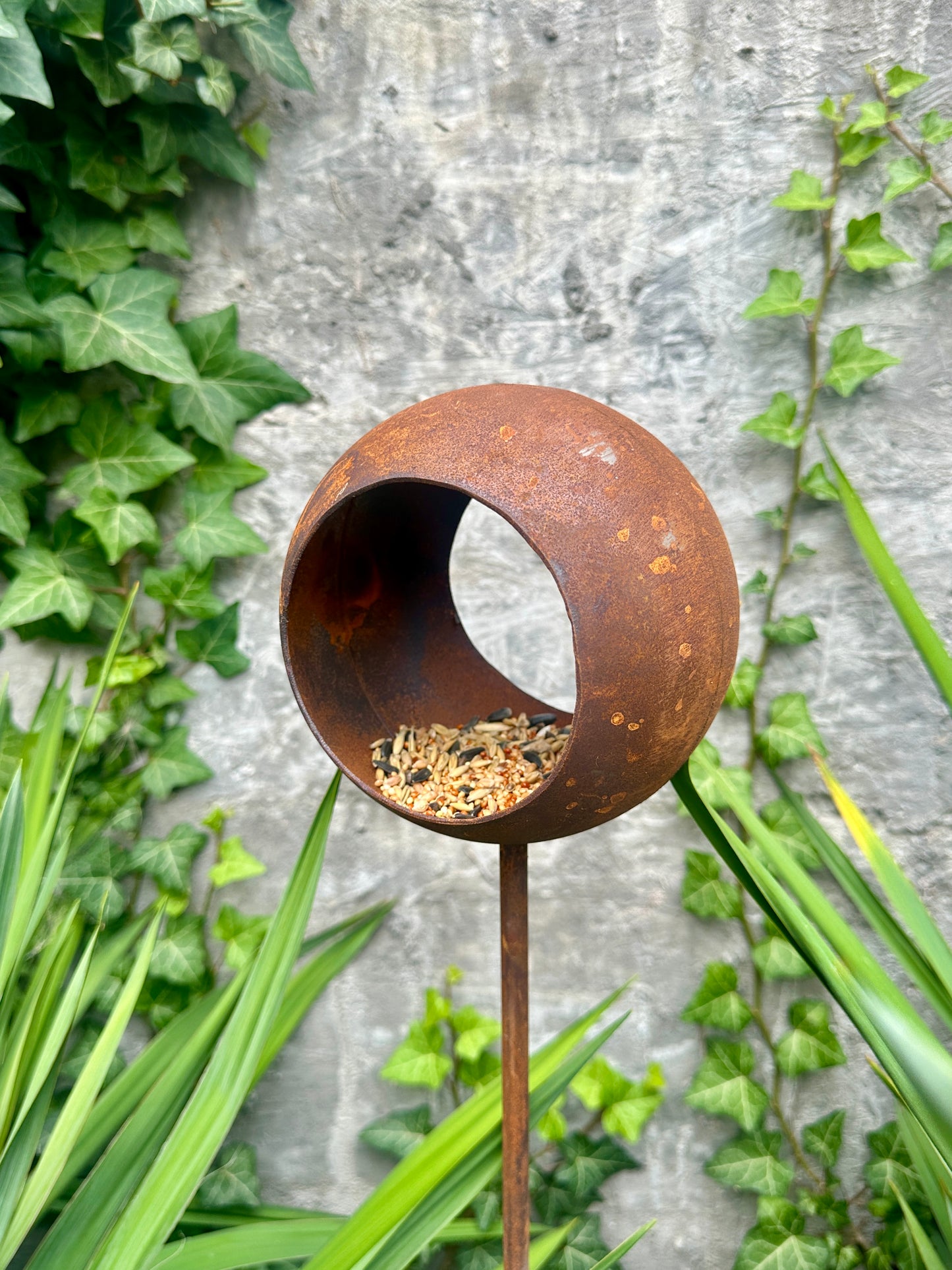 Rust-colored metal bird feeder on a stand with greenery in the background