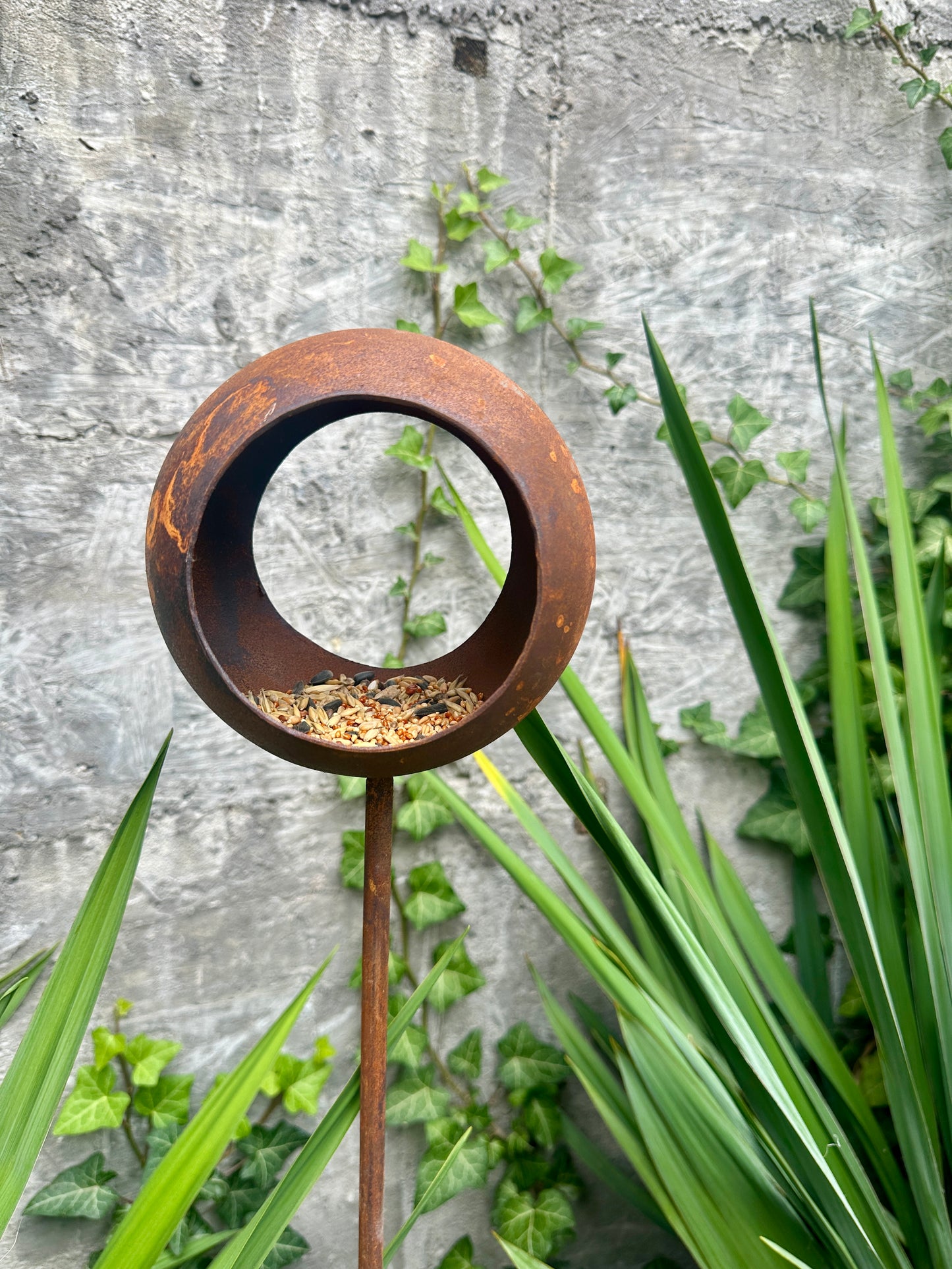 Rust-colored metal sculpture with a circular design in a garden setting.