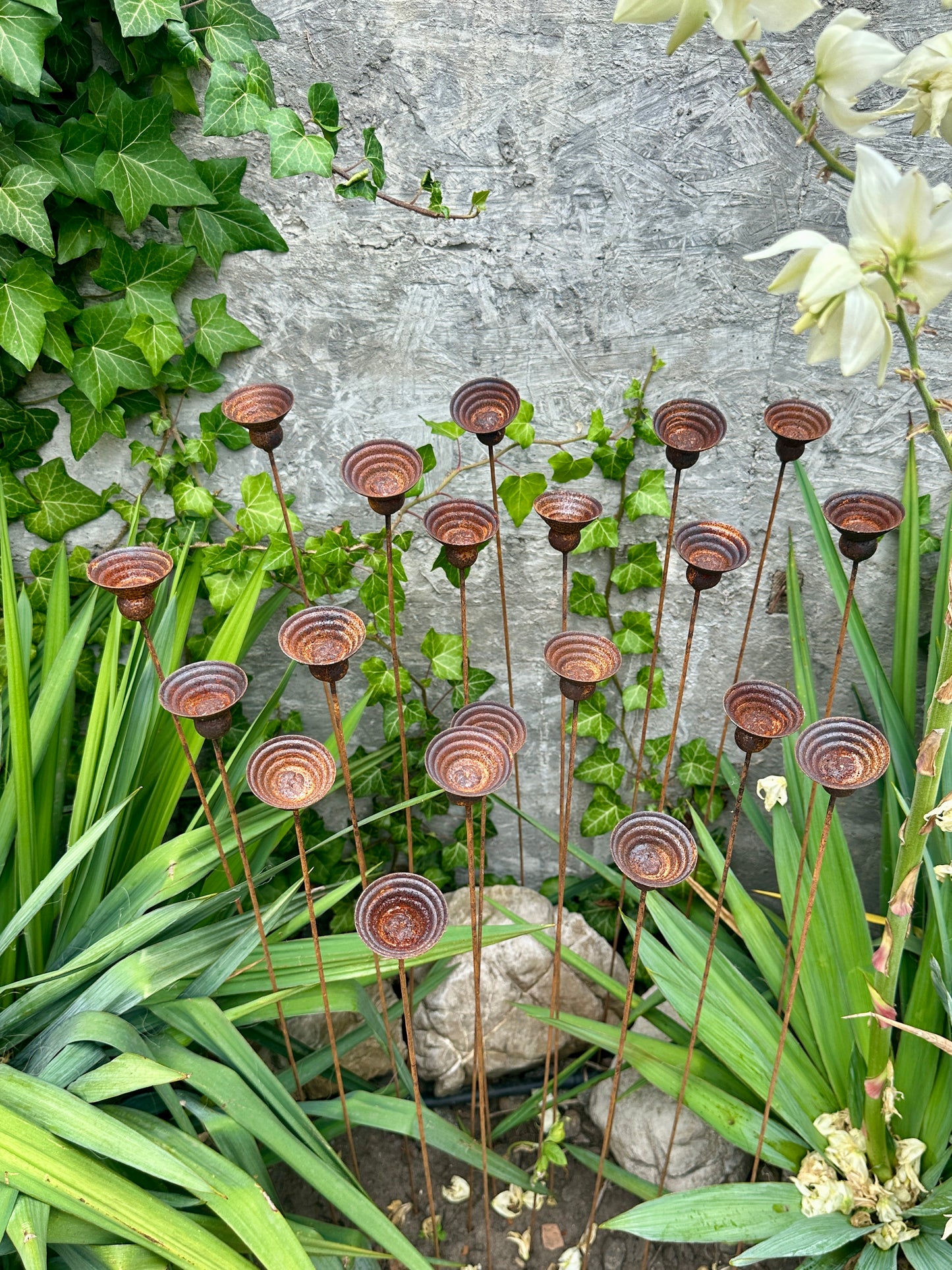 Decorative metal plant stands in a garden setting with greenery and a stone wall.