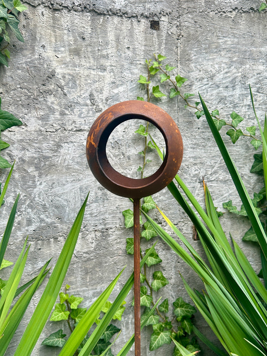 Decorative metal stake with circular design against a stone wall and greenery