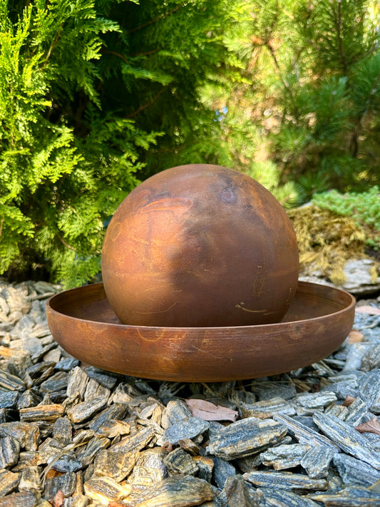 Rust-colored metal sphere and bowl on a stone surface with greenery in the background