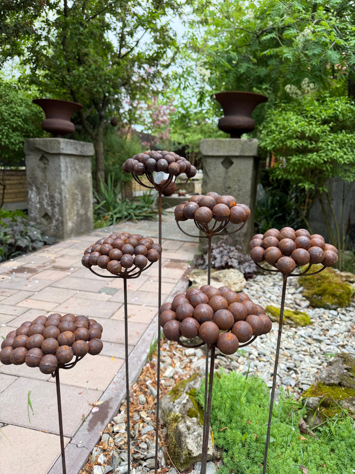 Decorative metal plant sculptures in a garden setting with trees and stone structures.