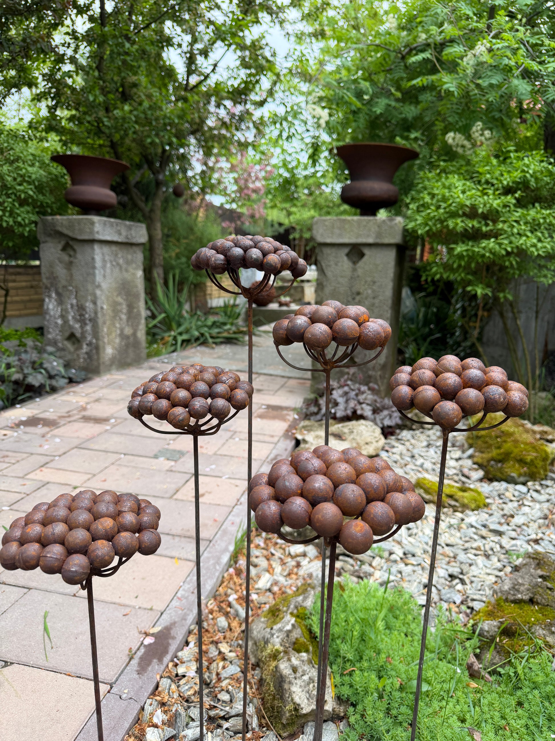 Decorative metal plant sculptures in a garden setting with trees and stone structures.