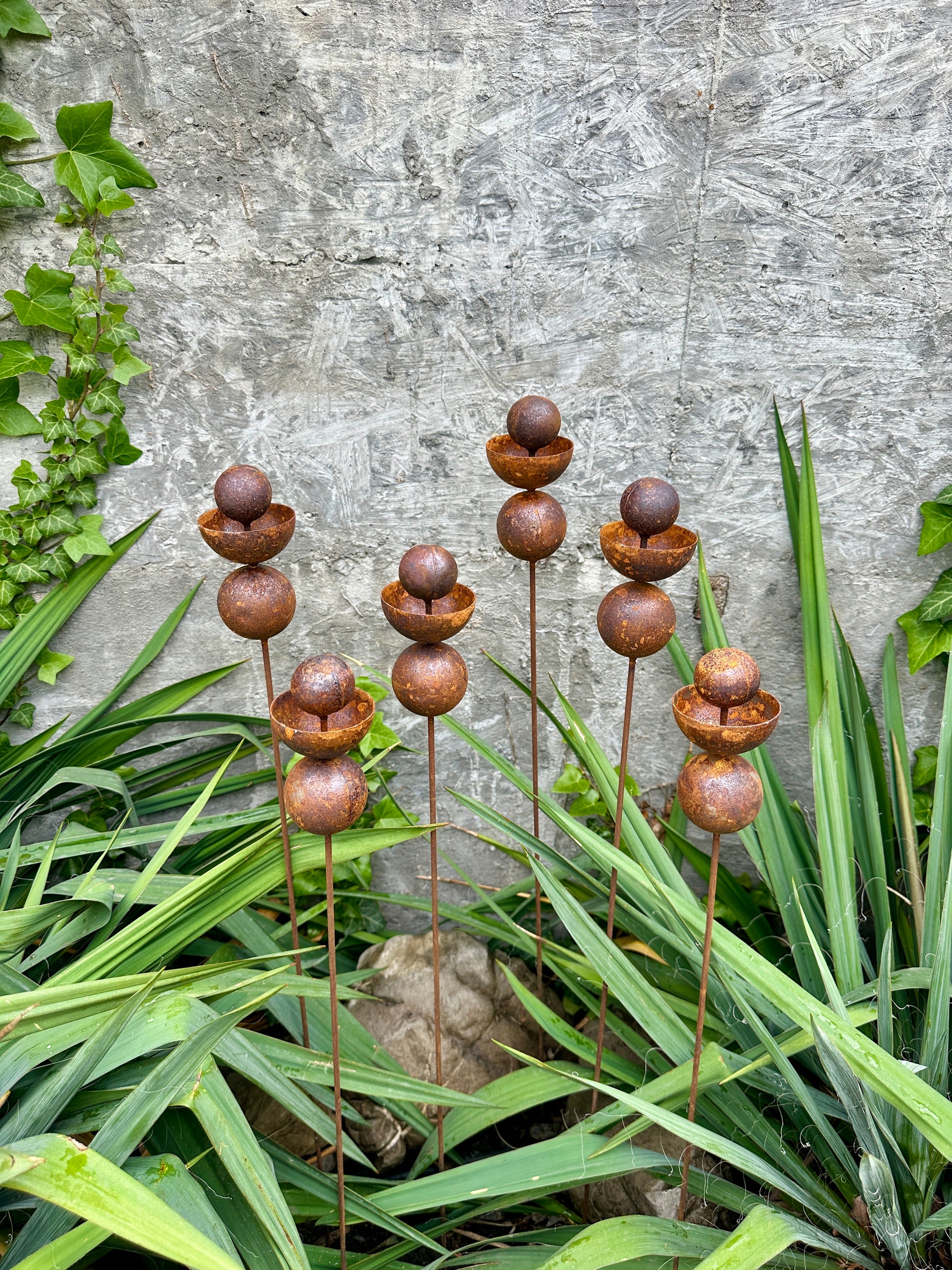 Decorative metal sculptures with rusted finishes on green plants against a stone wall.