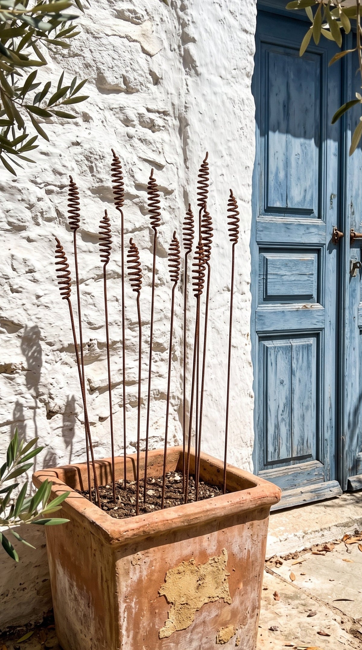 A terracotta square planter with dried decorative grass stems, placed outdoors against a white stucco wall next to a blue wooden door.