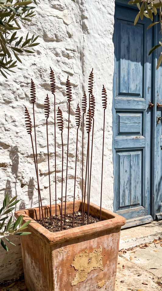 A terracotta square planter with dried decorative grass stems, placed outdoors against a white stucco wall next to a blue wooden door.